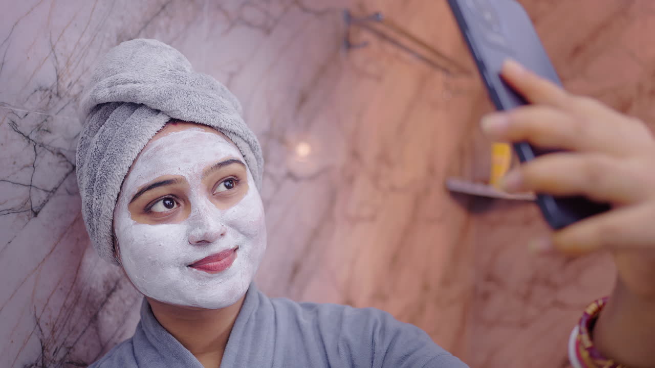 Donna indiana con una maschera facciale che si fa un selfie con il suo smartphone in un accogliente ambiente bagno.