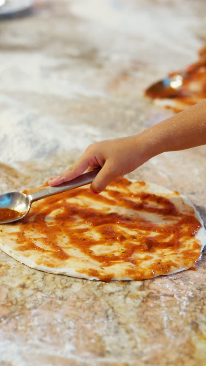 Vertical close-up of a little girl's hands using a spoon to spread tomato sauce on pizza dough during a fun cooking class