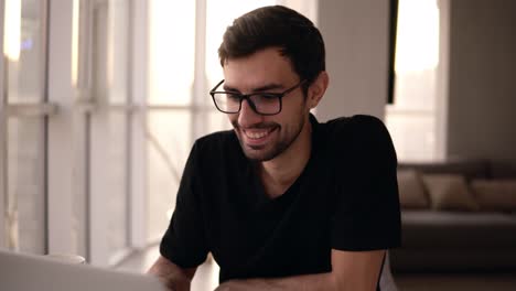 portrait of a toothy smiling man in glasses using laptop. freelancer working on computer at home. handsome man actively typing on laptop, chatting with friends at big house with panoramic windows and sofa on background