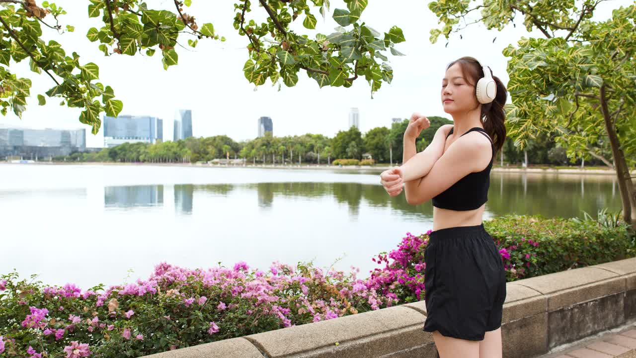 若い女性が湖畔で、背景に都市の建物を見ながら、腕のストレッチをしている。