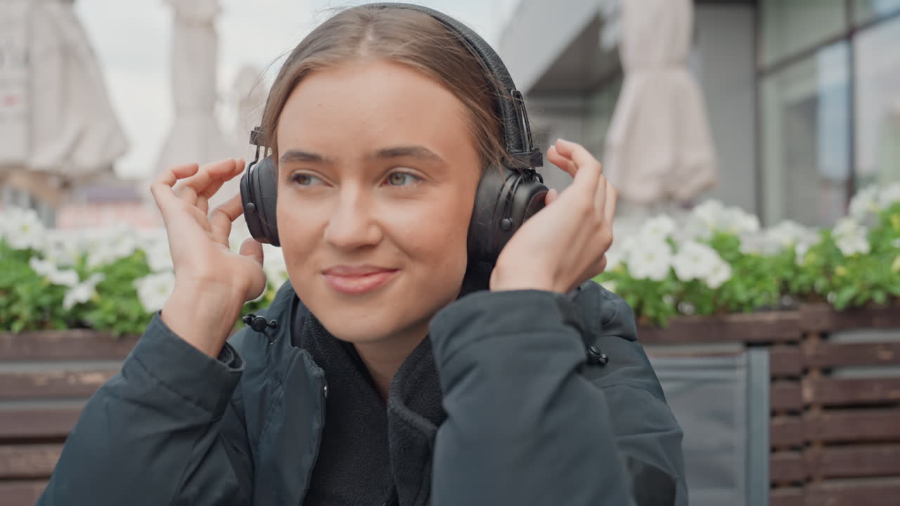 Giovane donna caucasica che ascolta musica, seduta al tavolino di un bar all'aperto con giacca nera, cuffie over-ear, occhi chiusi, che regola le cuffie, sorriso sereno, vasi di fiori, tavolo di vetro, sfondo urbano accogliente