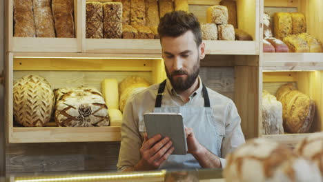 Junger-Mann-Brotverkäufer-Scrollt-Und-Klebt-Auf-Dem-Tablet-Computer,-Während-Er-An-Der-Theke-In-Der-Bäckerei-Steht,-Und-Schaut-Dann-In-Die-Kamera