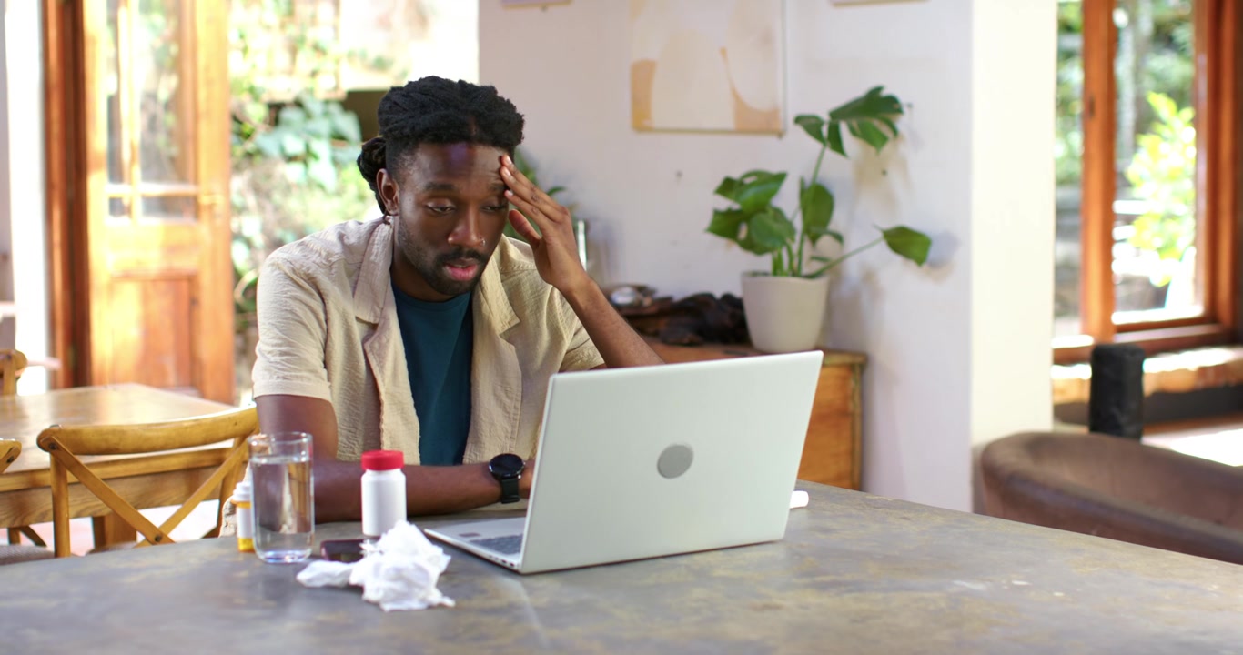 Homme afro-américain lisant un e-mail urgent sur son ordinateur portable à la maison, se frottant le front, avec un flacon de pilules, cherchant à se calmer.