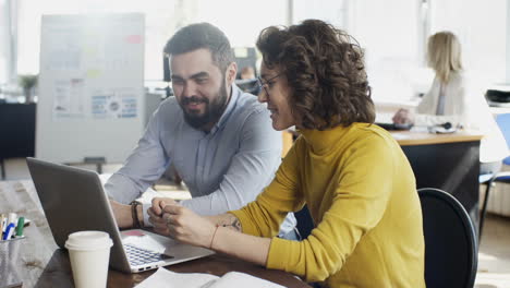 Joven-Alegre-Sentada-Colega-Masculino-En-El-Escritorio-De-La-Oficina,-Sonriendo-Y-Discutiendo-La-Presentación-En-La-Computadora-Portátil