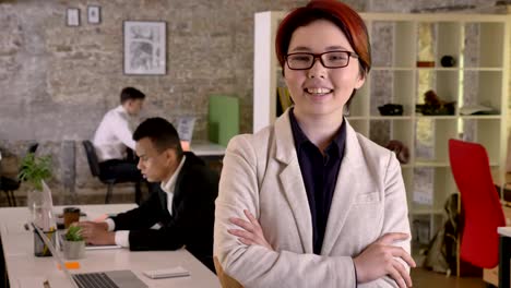 young business asian woman with crossed hands looking in camera in modern office and nodding, smiling, black and caucasian mans in back