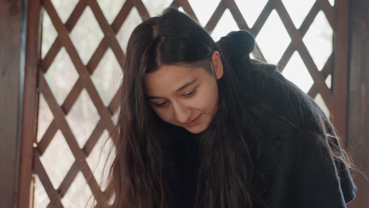Young woman leans forward inside wooden lattice shelter, downward gaze and gentle smile visible in soft daylight; dark hair frames face while calm focused expression suggests reflective mood and