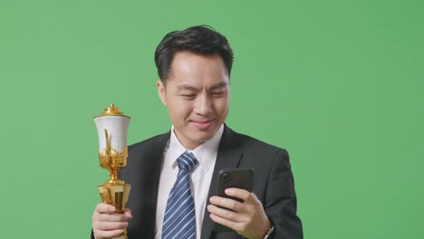 close up of asian business man in a suit and tie with a gold medal holding a gold trophy and using smartphone on green screen background in the studio