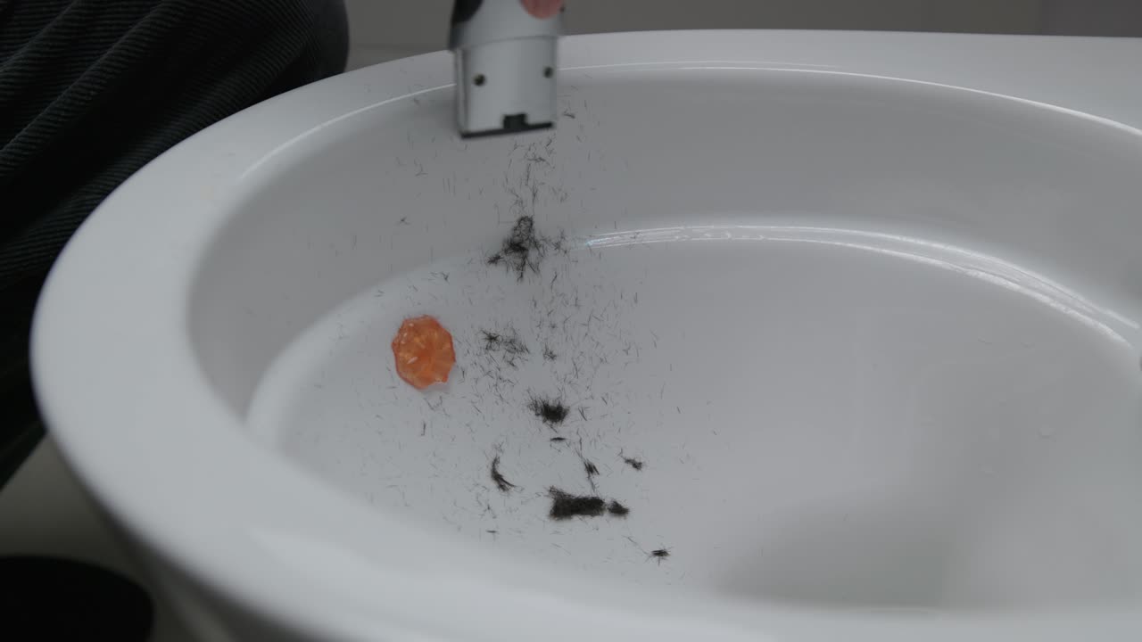 Side angle of a white toilet bowl with fresh hair clippings inside after shaving a head. A razor is tapped out again, small hairs falling into the bowl in a simple bathroom setting