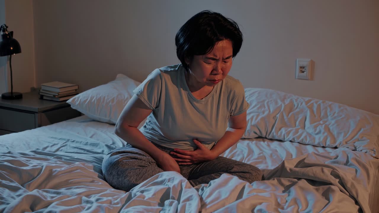 A woman sits on a bed in low light, holding her stomach in discomfort