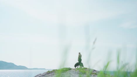 A-Woman-Hiker-With-Her-Dog-On-Leash-Standing-On-A-Rocky-Shore-Of-Donnamannen-Mountain-In-Nordland,-Norway