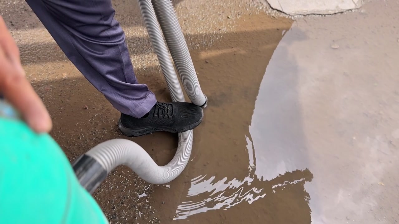 High-angle close-up of a worker using a heavy-duty wet vacuum hose to remove standing water from a puddle on asphalt. Professional equipment used for flood cleanup and outdoor maintenance.