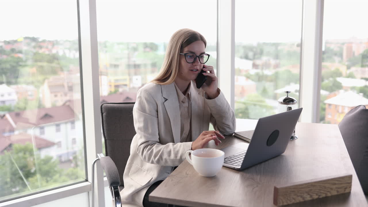 Bella giovane donna con gli occhiali che lavora in un ufficio luminoso e beve tè al laptop. Affascinante signora dai capelli lunghi con gli occhiali che parla al telefono