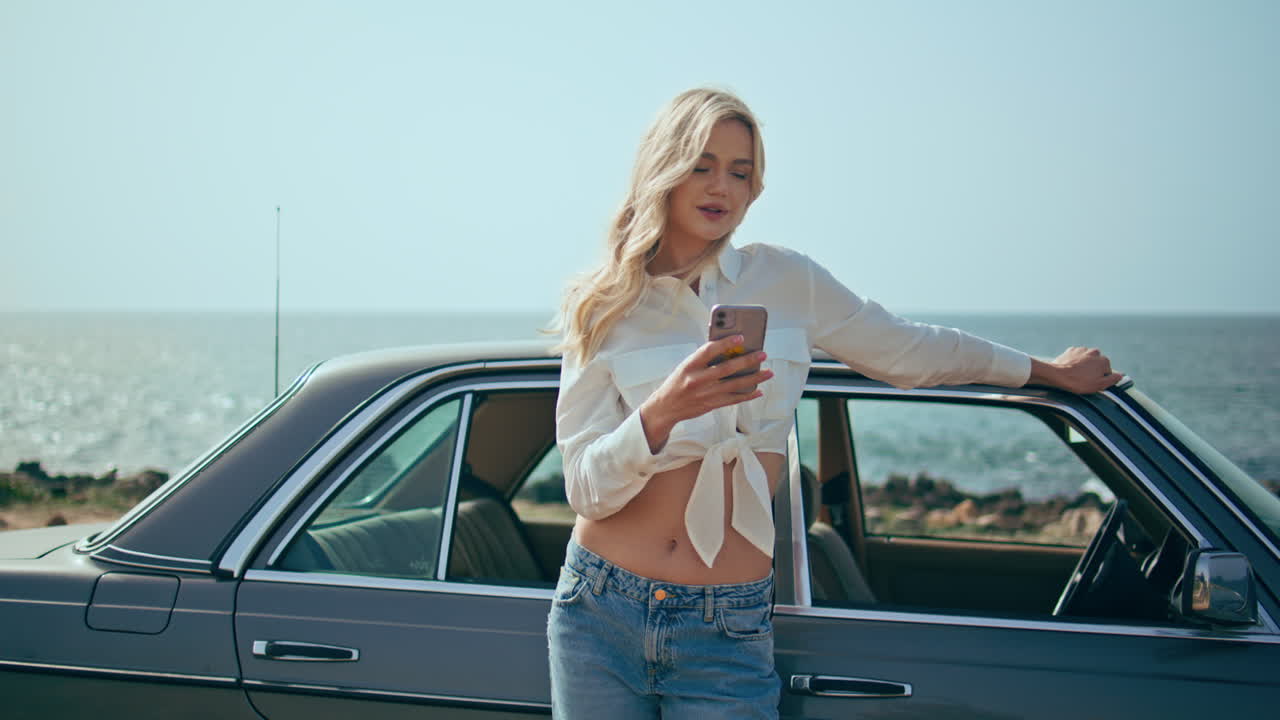 mujer termina una llamada telefónica en la costa soleada apoyándose en un automóvil retro. viaje marítimo