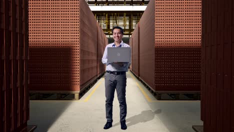 full body of an asian male professional worker standing with his laptop with red brick packed in stacks are stored, he is looking at the camera with a smile