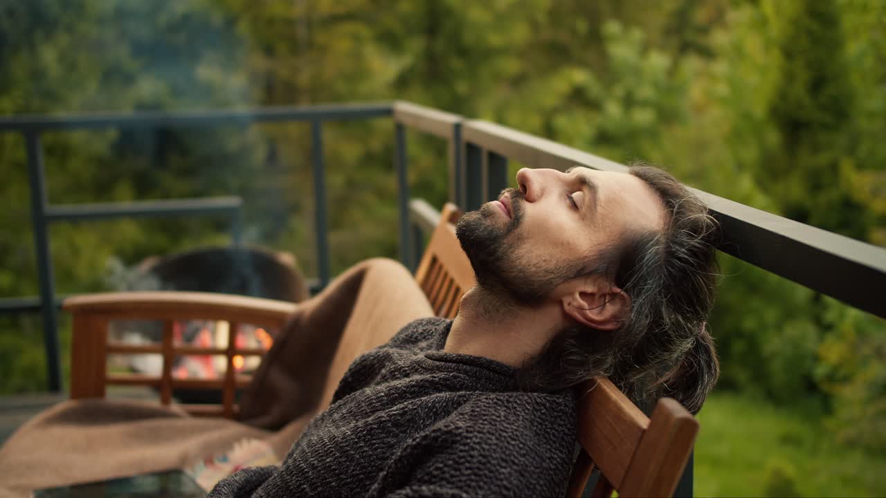 un joven moreno con los ojos cerrados descansa en un sofá en el balcón de una casa de campo con vistas a las montañas y un bosque de coníferas, la leña se quema cerca de él en la barbacoa