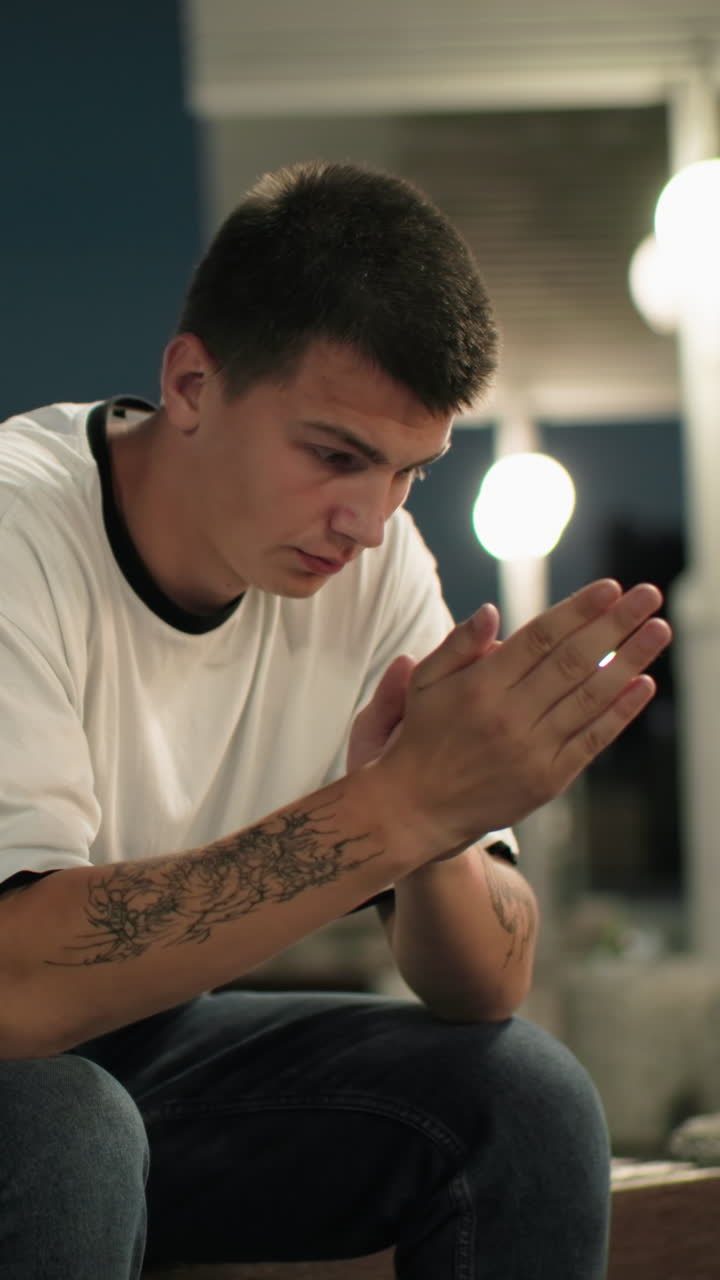 Contemplative man with tattooed hand seated on bench at dusk, deep in thought, urban background with streetlights and blurred couples passing by