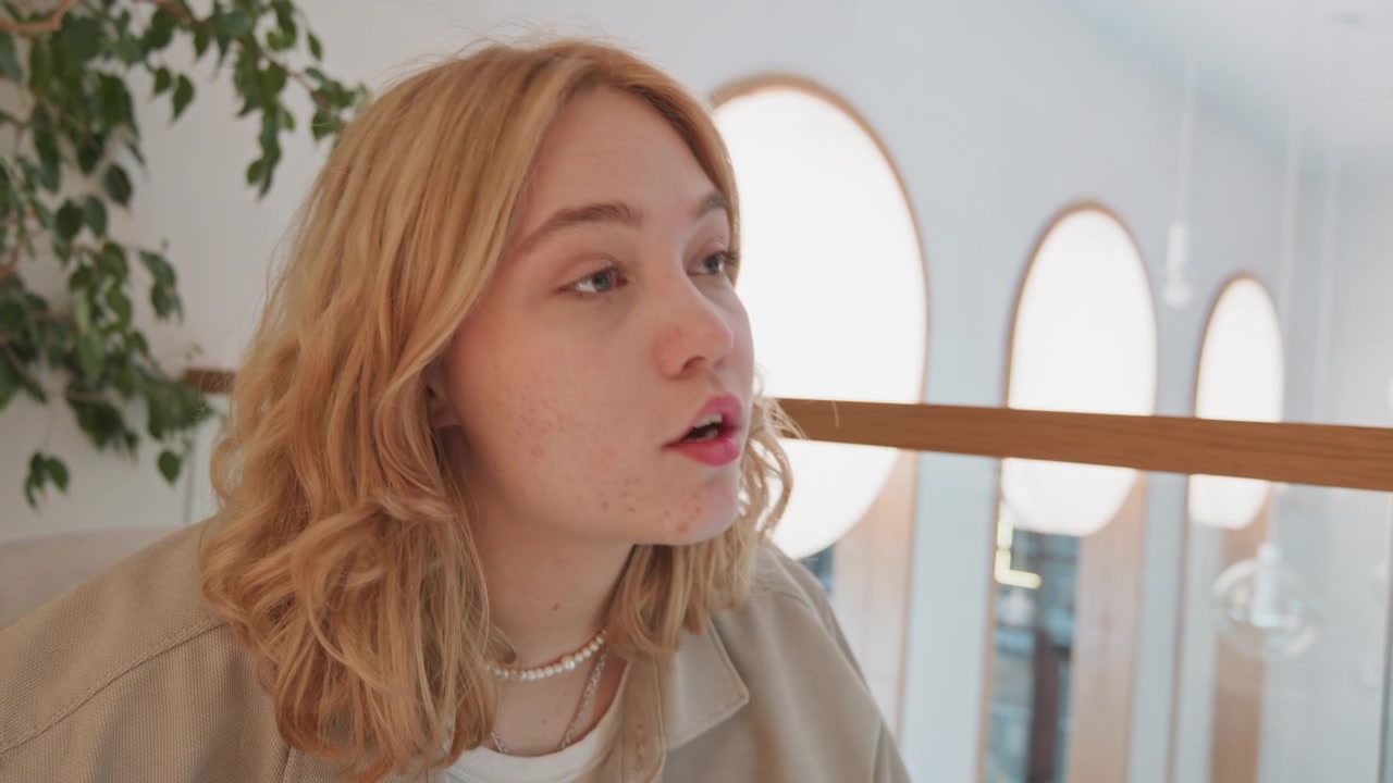 Close up of young woman with wavy blonde hair wearing layered necklaces and beige jacket seated in indoor setting. scene features soft natural light streaming through large round windows