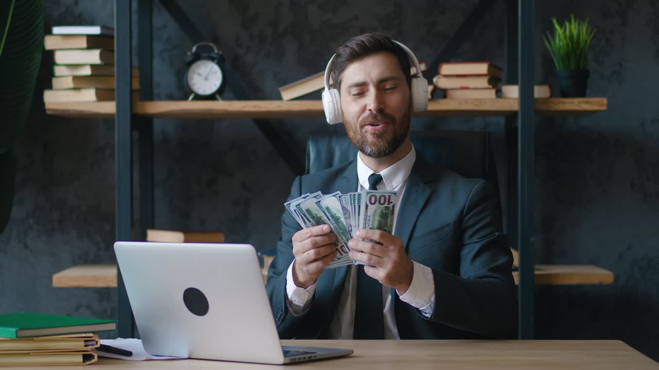 счастливый бородатый молодой бизнесмен слушает музыку на наушниках и любит считать деньги, долларовые купюры. концерт легкой прибыли