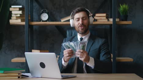 feliz joven empresario barbudo escucha música en auriculares y disfruta contando dinero, billetes de dólar. concierto de beneficio fácil