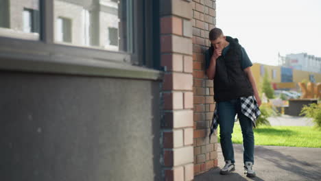 young male wearing puffer vest and checkered shirt tied around waist leans against brick wall while inhaling vape device and coughing on urban street with modern buildings in bright natural light