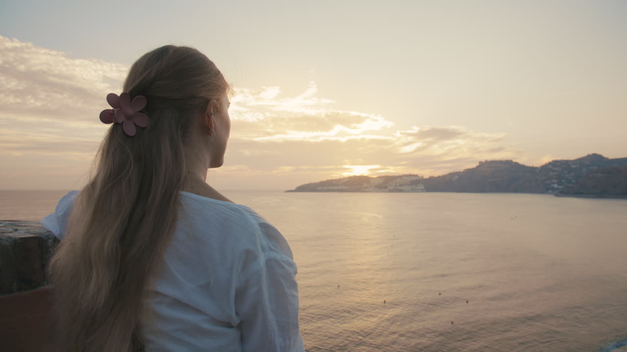 Medium over-the-shoulder shot with soft focus depth, warm sunset light, and a calm ocean view—evoking reflection, peace, and quiet solitude