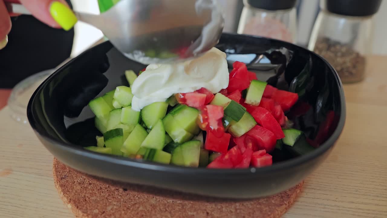 Woman adds sour cream with spoon in slow motion on fresh cucumber tomato salad