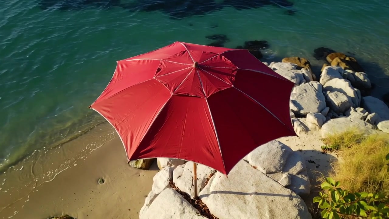 La telecamera inizia a scendere e a panoramare, rivelando un ombrello rosso su una sporgenza in una cala rocciosa.