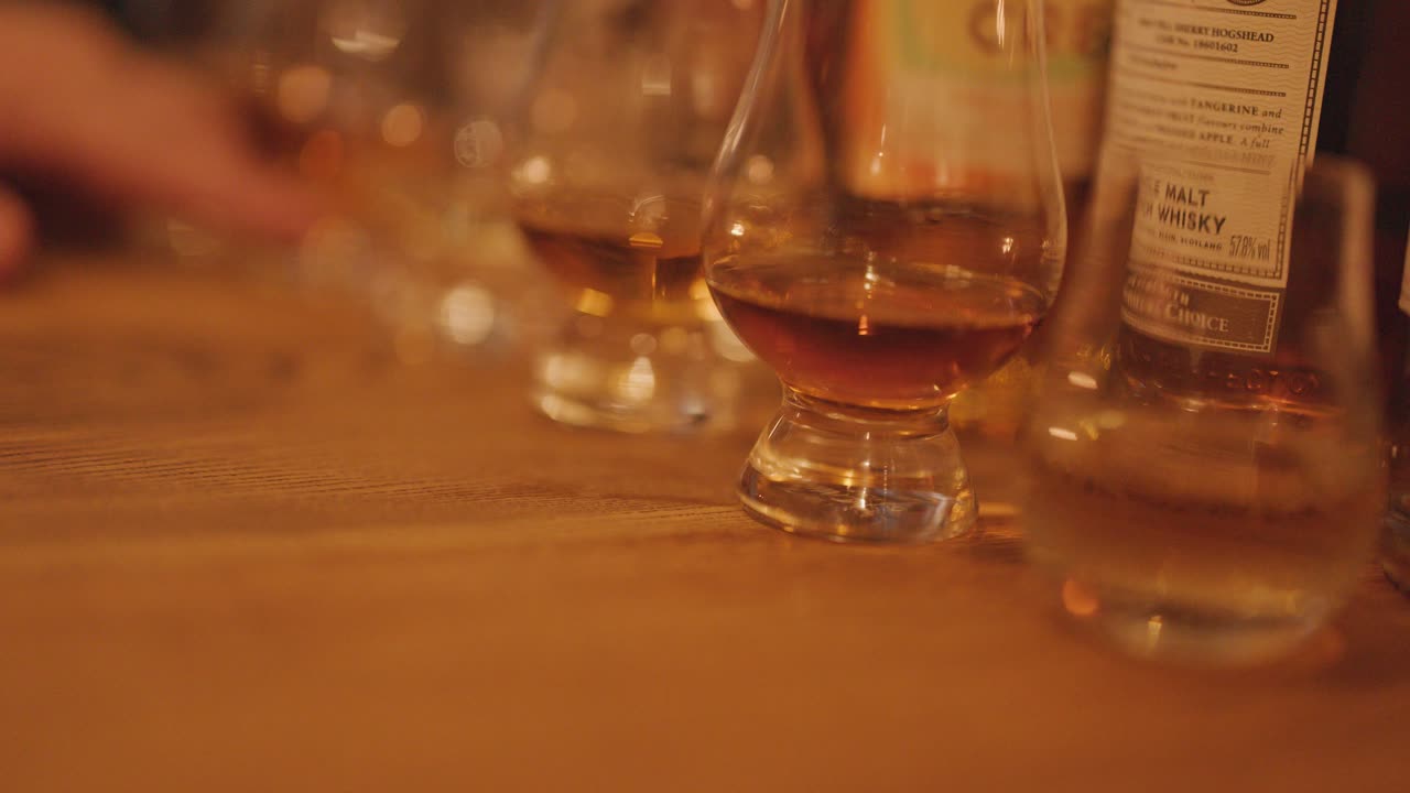 A hand reaches for a whisky glass on a wooden table, surrounded by tasting glasses and bottles in soft, ambient lighting with shallow depth of field
