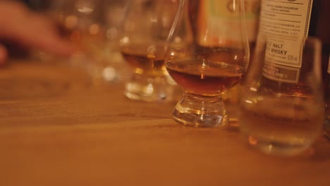 a hand reaches for a whisky glass on a wooden table, surrounded by tasting glasses and bottles in soft, ambient lighting with shallow depth of field