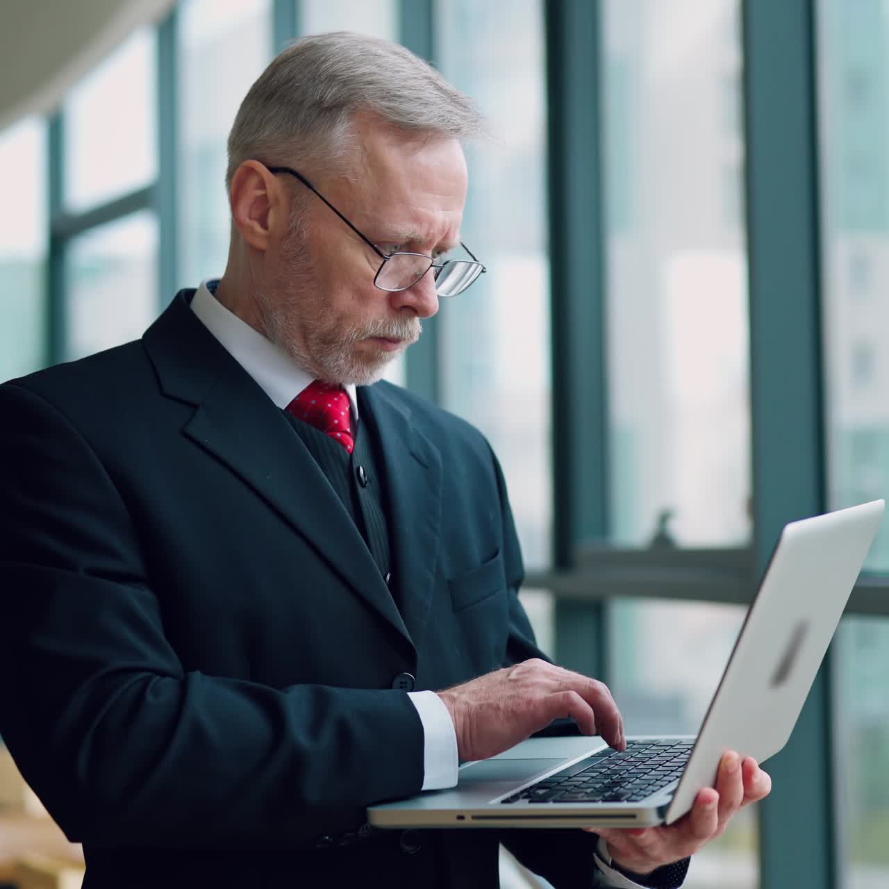 Successful businessman in glasses with a laptop in the office. Elegant mature man in suit standing near the window and holds a modern laptop.