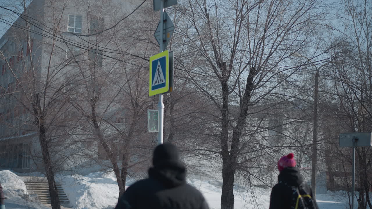 winter street lined with snow piles and bare trees, urban building backdrop, yellow pedestrian sign with countdown, students walking along icy sidewalk in cold daylight beneath clear sky