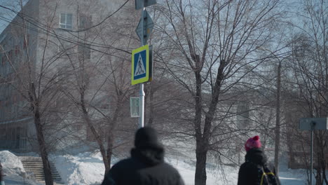 winter street lined with snow piles and bare trees, urban building backdrop, yellow pedestrian sign with countdown, students walking along icy sidewalk in cold daylight beneath clear sky