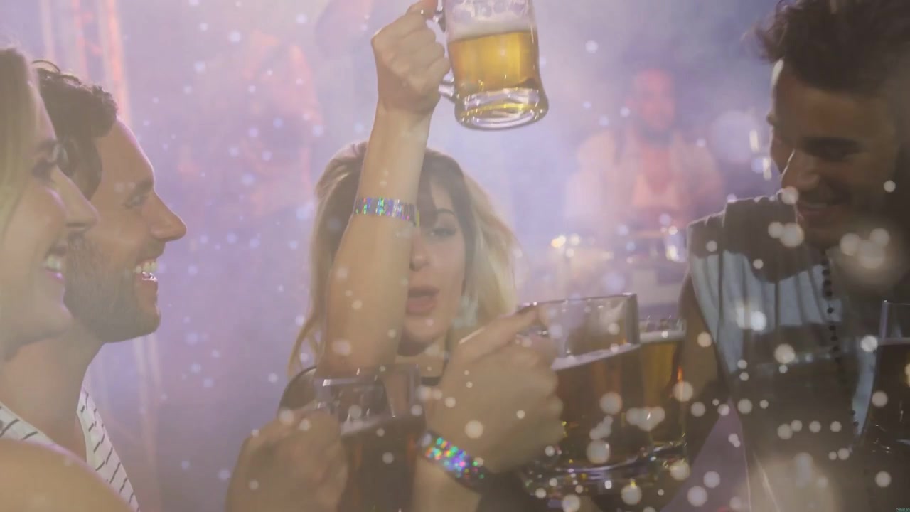 Fünf Freunde stoßen im Club mit Tassen an, während ein funkelndes Konfetti-Overlay zu sehen ist und sie sich vorbeugen, um auf ein Telefon zu schauen.