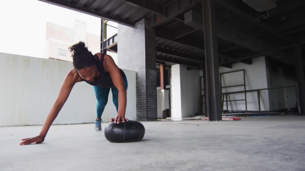 mujer afroamericana haciendo ejercicios de flexión en una pelota de medicina en un edificio urbano vacío