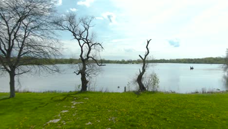 a low to the ground flight through a couple trees onto an open space above a small lake in a small national park in a suburban area