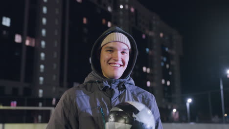 jovem adulto vestindo capuz segurando uma bola de futebol na mão e rindo alegremente enquanto olhava para a câmera durante a noite em um campo de esportes urbano com edifícios de escritórios iluminados e luzes de rua