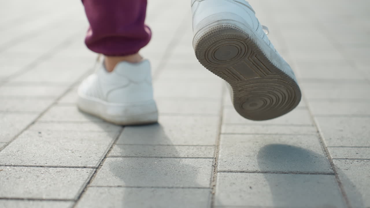 Back leg view of female athlete walking on gray pavement under sunny sky casting long shadows capturing sneaker detail and confident stride in urban sidewalk with morning light and casual outfit