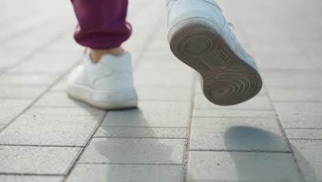 back leg view of female athlete walking on gray pavement under sunny sky casting long shadows capturing sneaker detail and confident stride in urban sidewalk with morning light and casual outfit