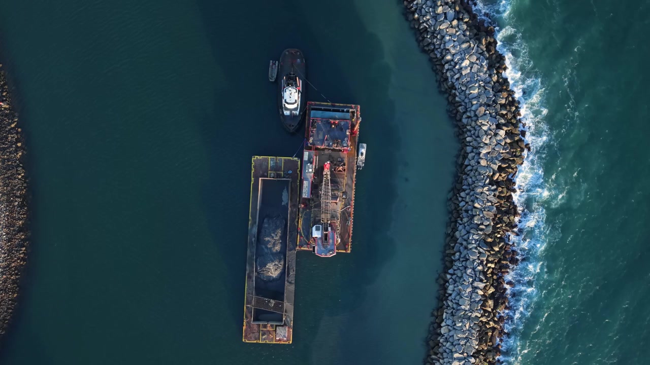Industrial barge and tugboat float beside a rocky harbor breakwater in blue coastal water with dreding equipment, aerial top-down orbit