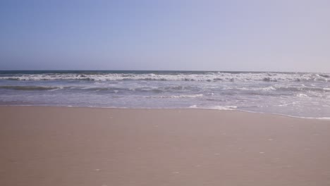 Low-level-pan-shot-from-sand-perspective-showing-waves-crashing-onto-the-shore-in-Santa-Monica,-CA