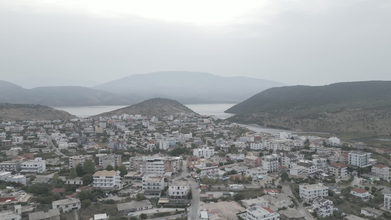 un avión no tripulado volando sobre ksamil albania por la mañana en un día gris nublado con niebla en el cielo y hoteles y casas blancas en el fondo con montañas y el registro del mar