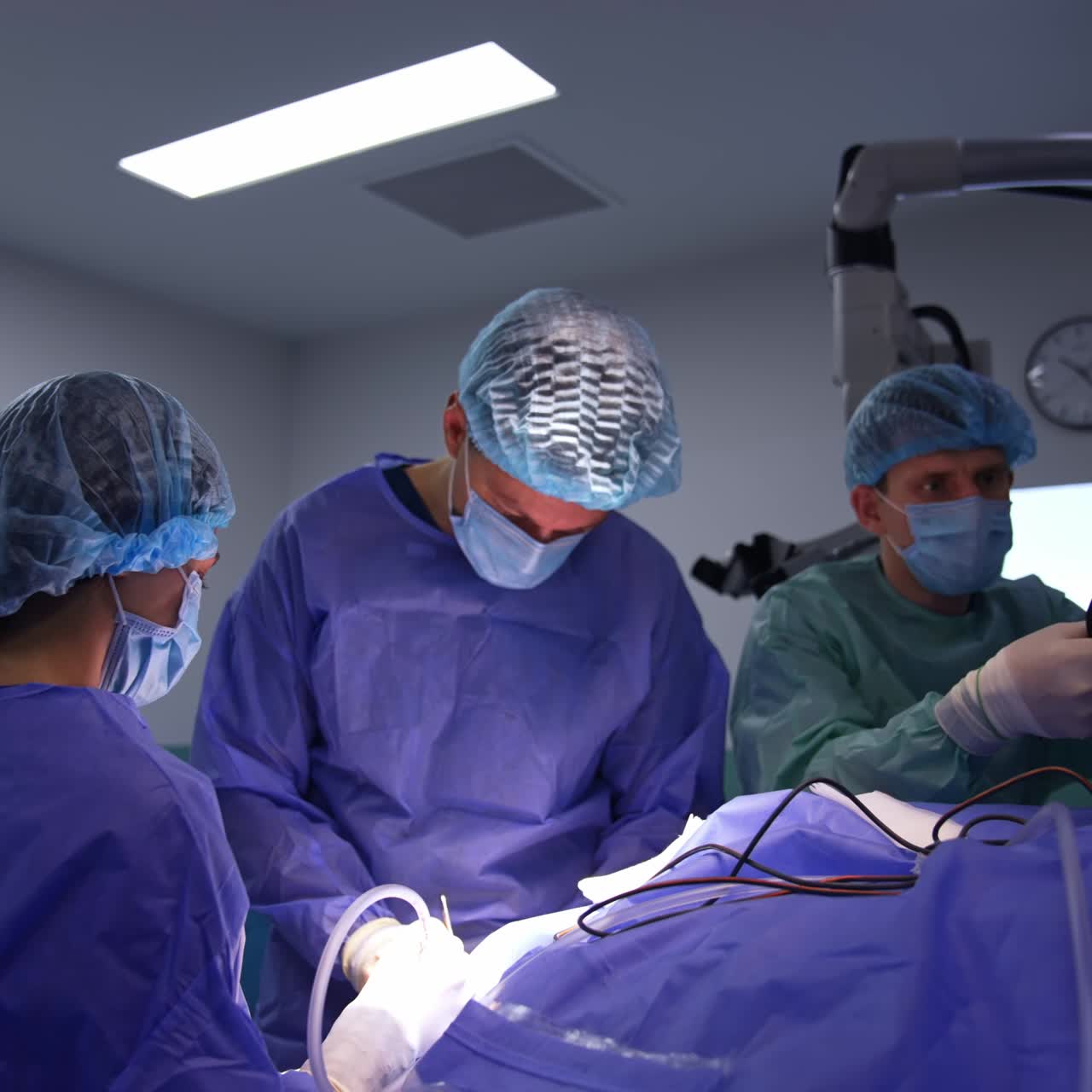 Surgeon conducts the operation using tools assisted by female nurse. Male assistant unties the wires of electric device