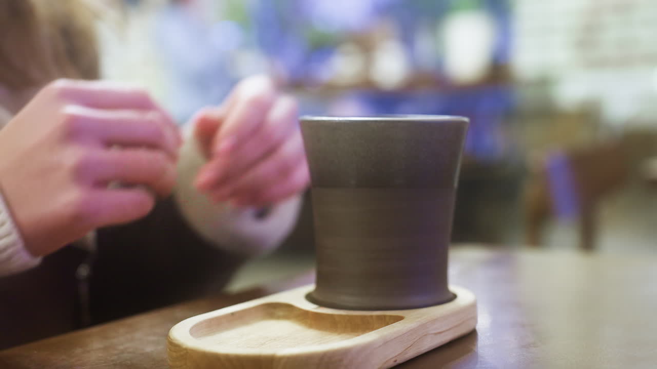 Close-up of woman's hand in brown shearling jacket, gently lifting cup of coffee at cozy cafe table. Soft lighting creates warm atmosphere, capturing peaceful moment of relaxation and enjoyment