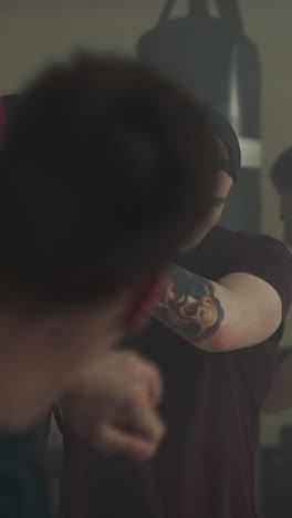 entrenador profesional con tatuaje en el brazo sostiene equipo de boxeo para que el estudiante practique kickboxing en un gimnasio oscuro. club de lucha para practicar tipos agresivos de deportes con el entrenador