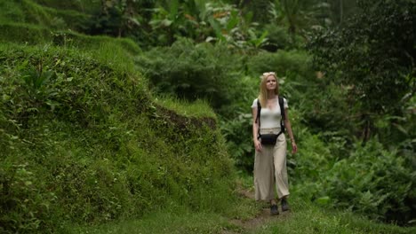 Attraktive-Junge-Blonde-Frau,-Die-Auf-Der-Reisterrasse-Und-Den-Reisfeldern-Auf-Die-Kamera-Zugeht-Und-Sich-Umsieht-Und-Die-Umliegende-Grüne-Umgebung-In-Ubud,-Bali,-Indonesien,-Genießt