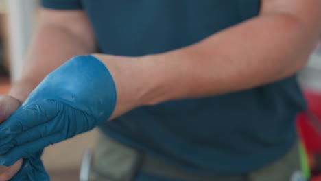 close up of man pulling blue disposable gloves off hand and dropping gloves onto workbench, tile wall background out of focus, red container blurred in frame, focusing on hand movement