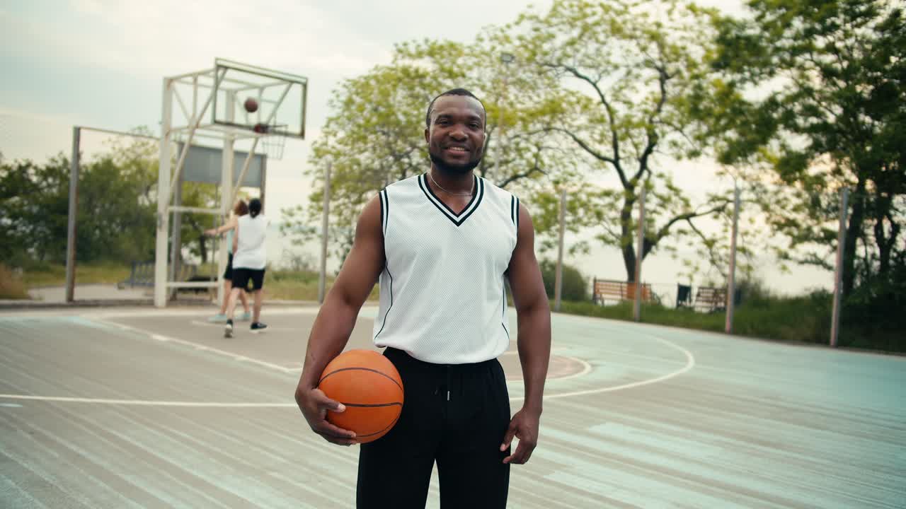 A black person basketball player with a basketball in his hands poses and looks at the camera against the background of his friends who are playing basketball on the street