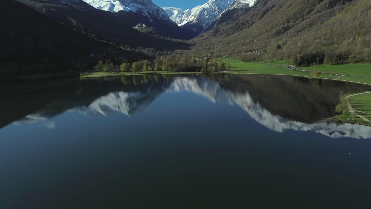 Snow capped mountains mirroring on Lake Génos-Loudenvielle in the French Pyrenees, aerial view with dolly backward and tilt up.