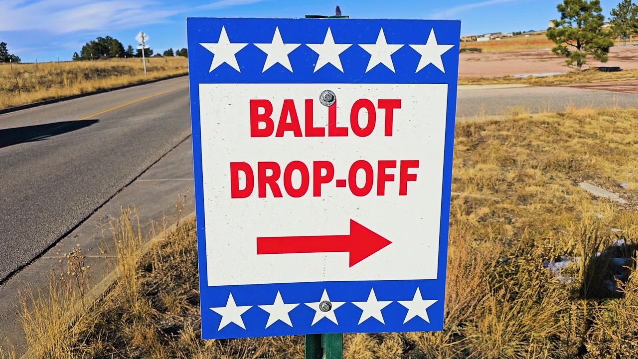 Patriotic ballot drop-off sign stands along a roadside with bold red text, white stars, and directional arrow, marking a voting location during a United States election season outdoors.