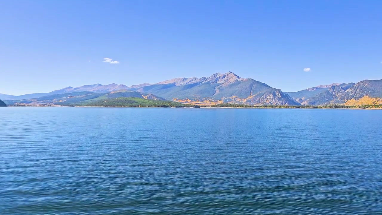秋のディロン湖の水面越しに広がる穏やかな眺め。水面にはさざ波が立ち、遠くには湖畔の家々、黄色いアスペンの木立、常緑樹の森、そして広大な山の景色が見える。